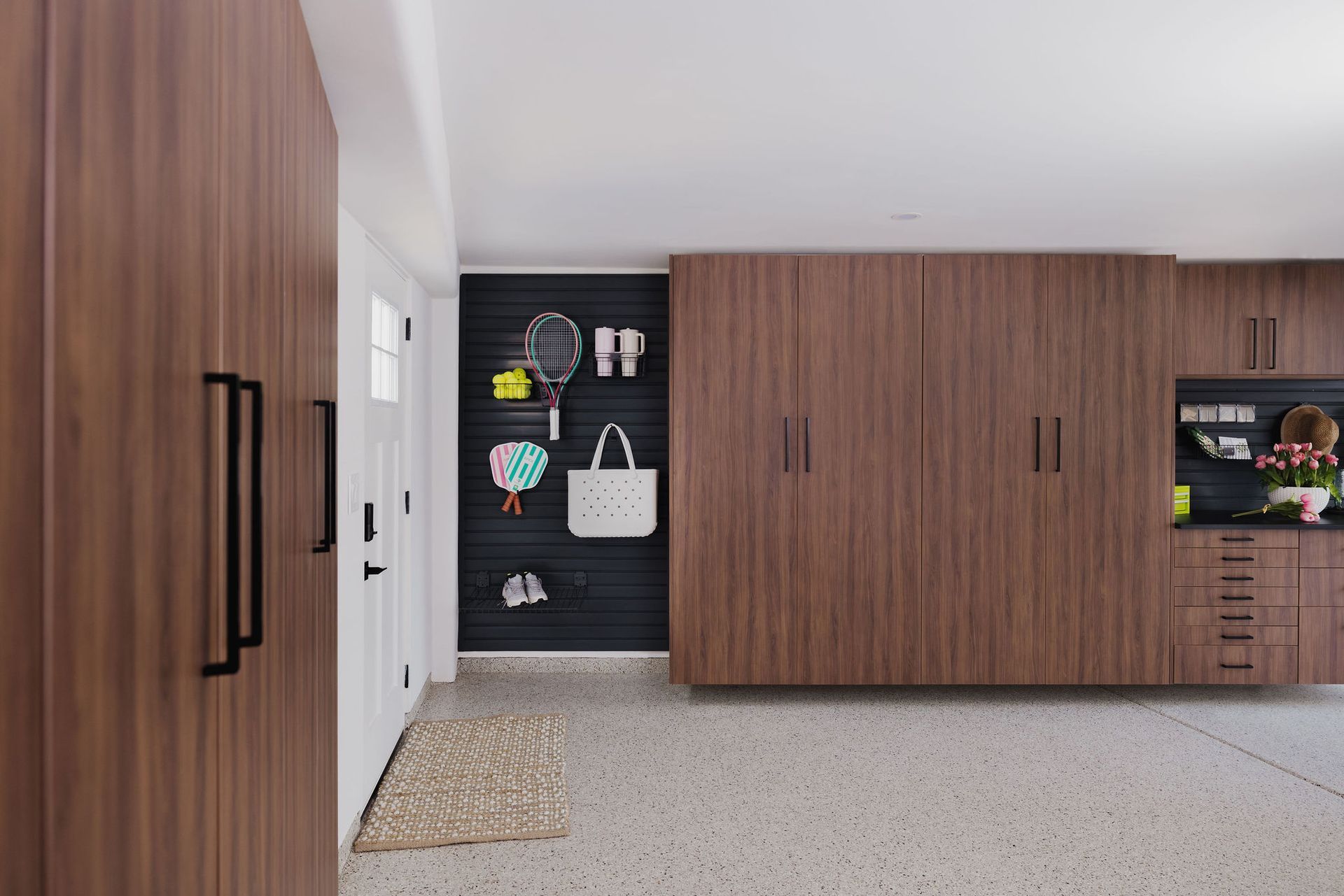 Modern organized garage with walnut cabinetry, a black slatted wall for hanging gear, and light terrazzo flooring.