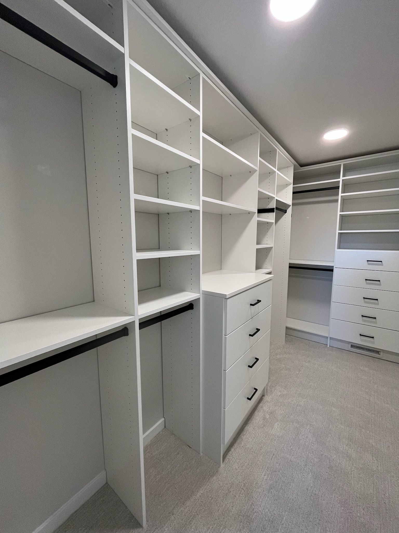 Walk-in closet with white shelving, drawers, and hanging rods on a carpeted floor
