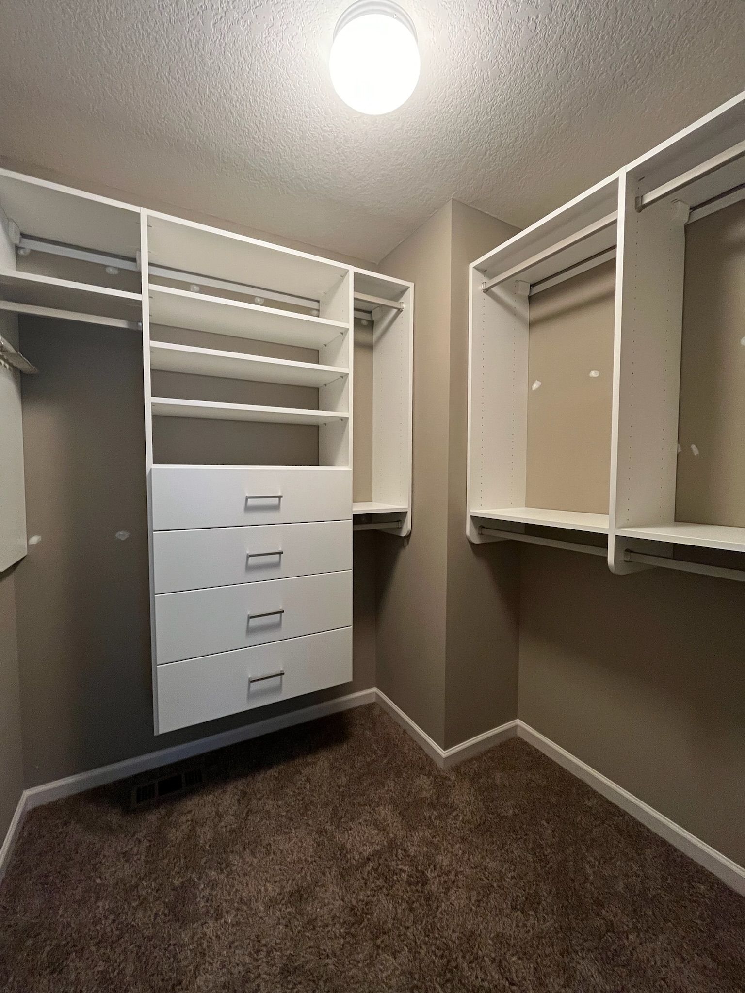 Walk-in closet with white shelving and drawers, beige walls, and brown carpet.