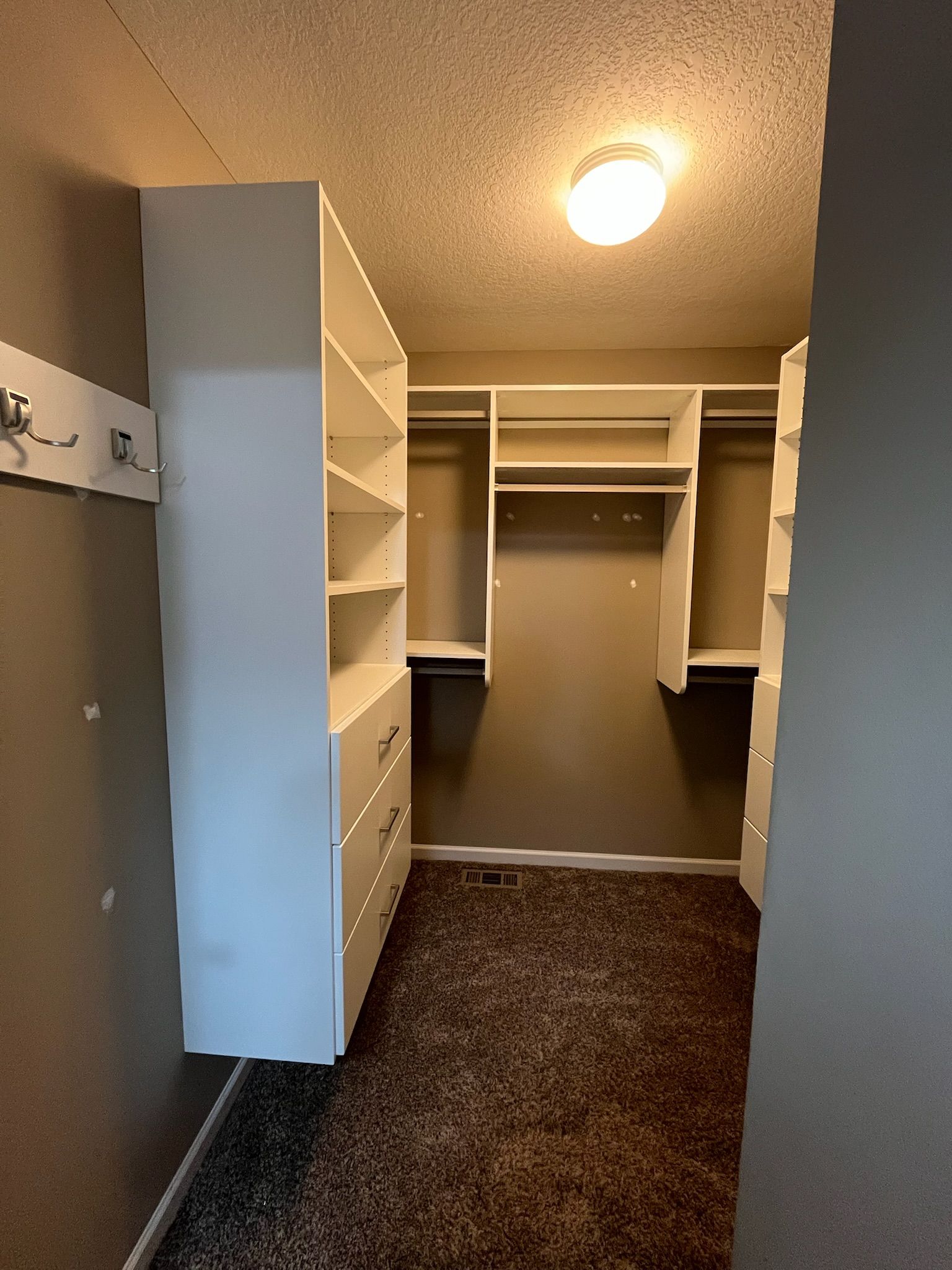 Empty walk-in closet with white shelving, drawers, and dark carpet under a ceiling light.