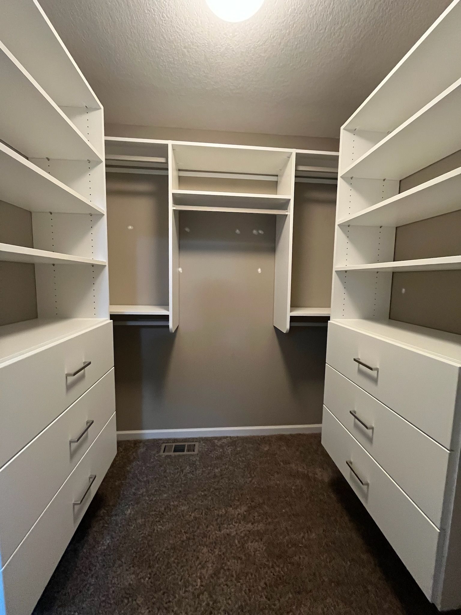 Walk-in closet with white shelves and drawers on both sides and a gray carpeted floor.