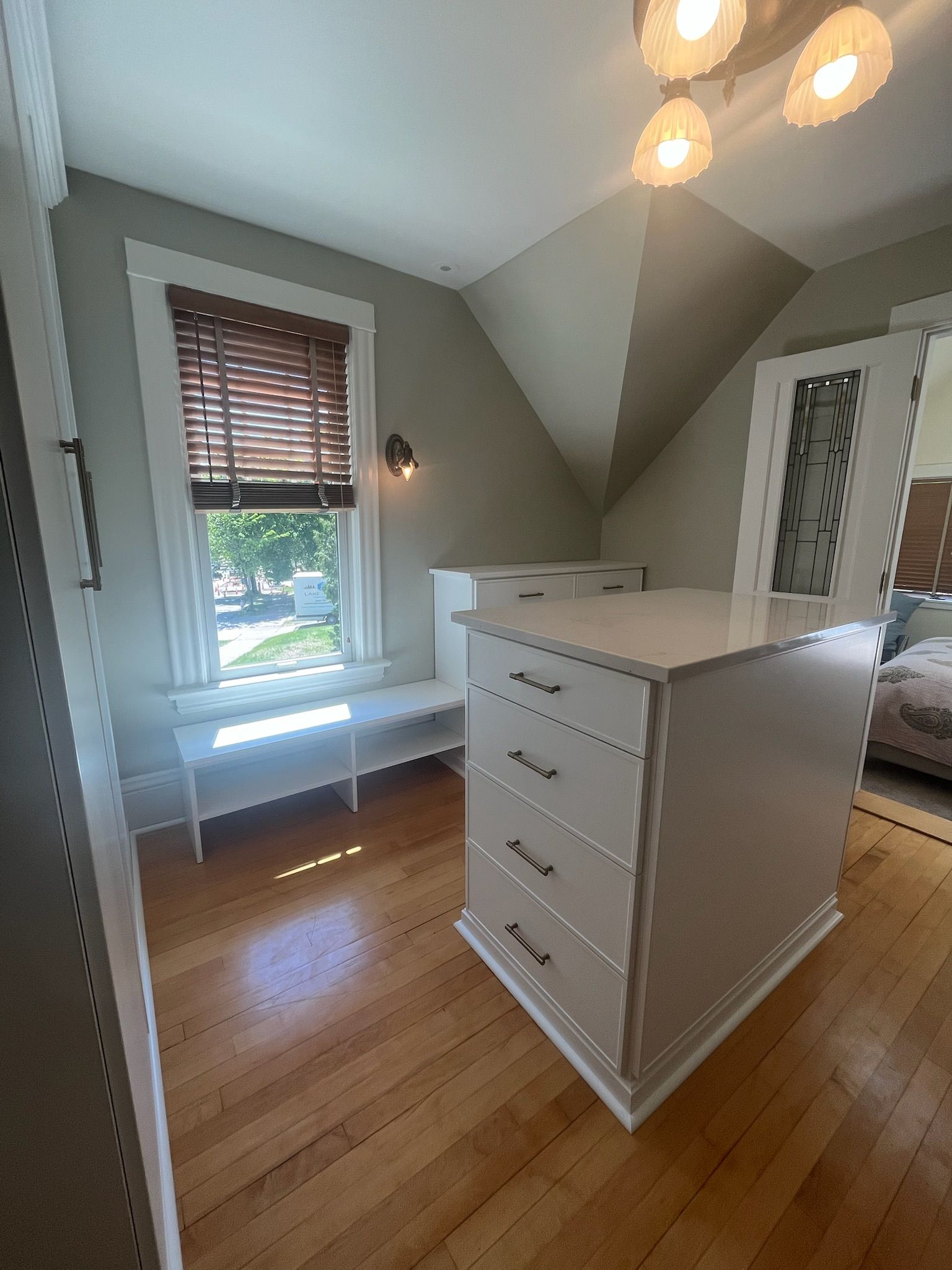 Bright bedroom with white dresser, bench by window, hardwood floors, and sloped ceiling lights