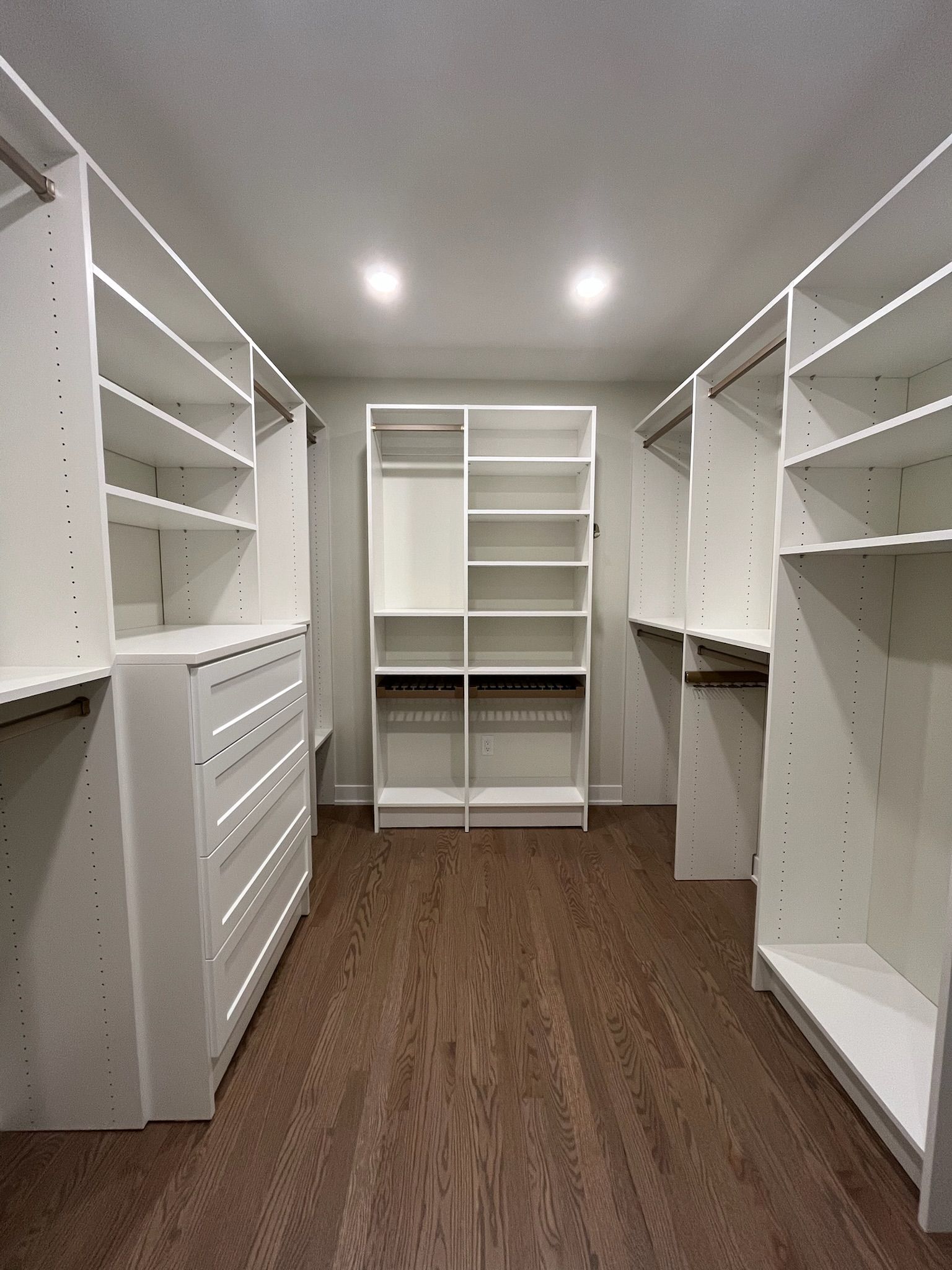 Empty walk-in closet with white shelving and drawers, wood floor, and ceiling lights.