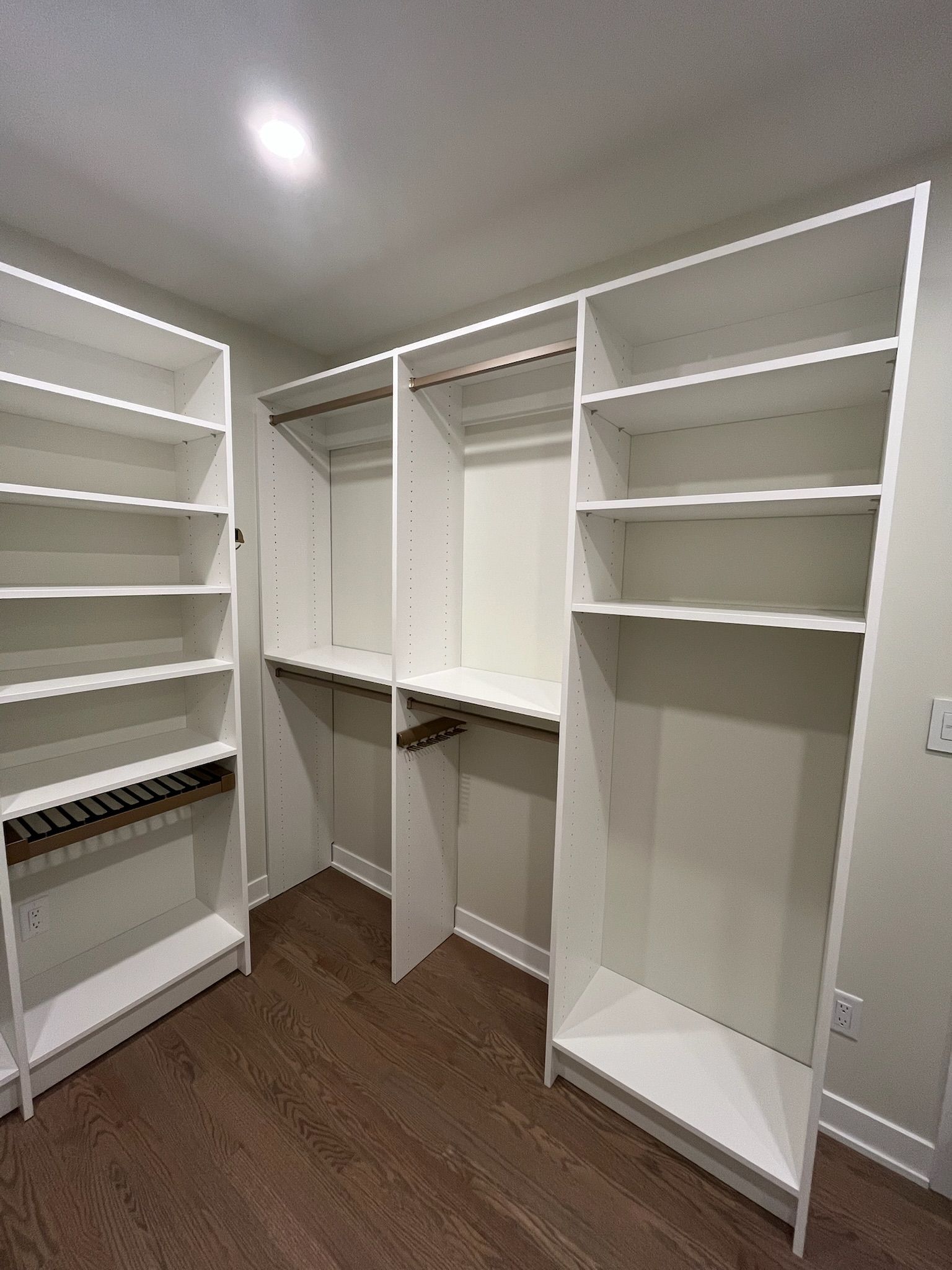 Empty walk-in closet with white shelves, hanging rods, and brown wood flooring.