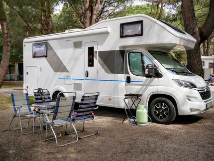 White RV camper parked at a wooded campsite with folding chairs, table, and grill outside