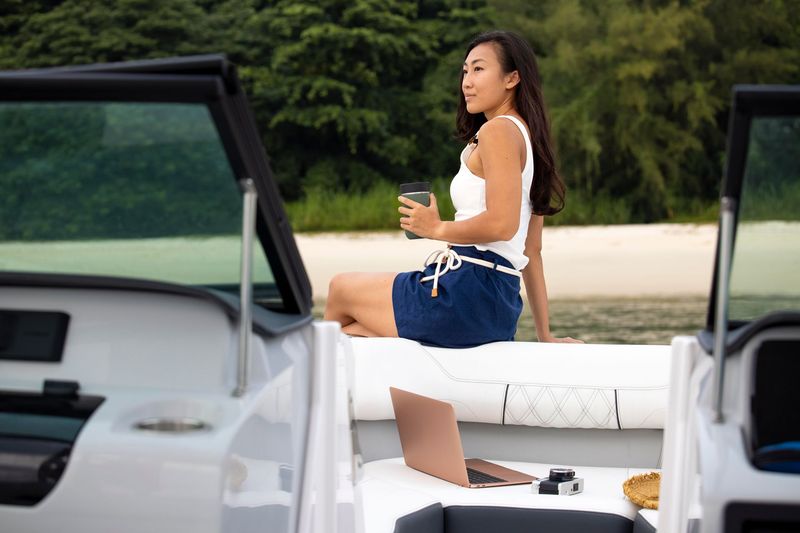 Person sitting on a white boat seat holding a drink, with open boat doors and trees in the background.