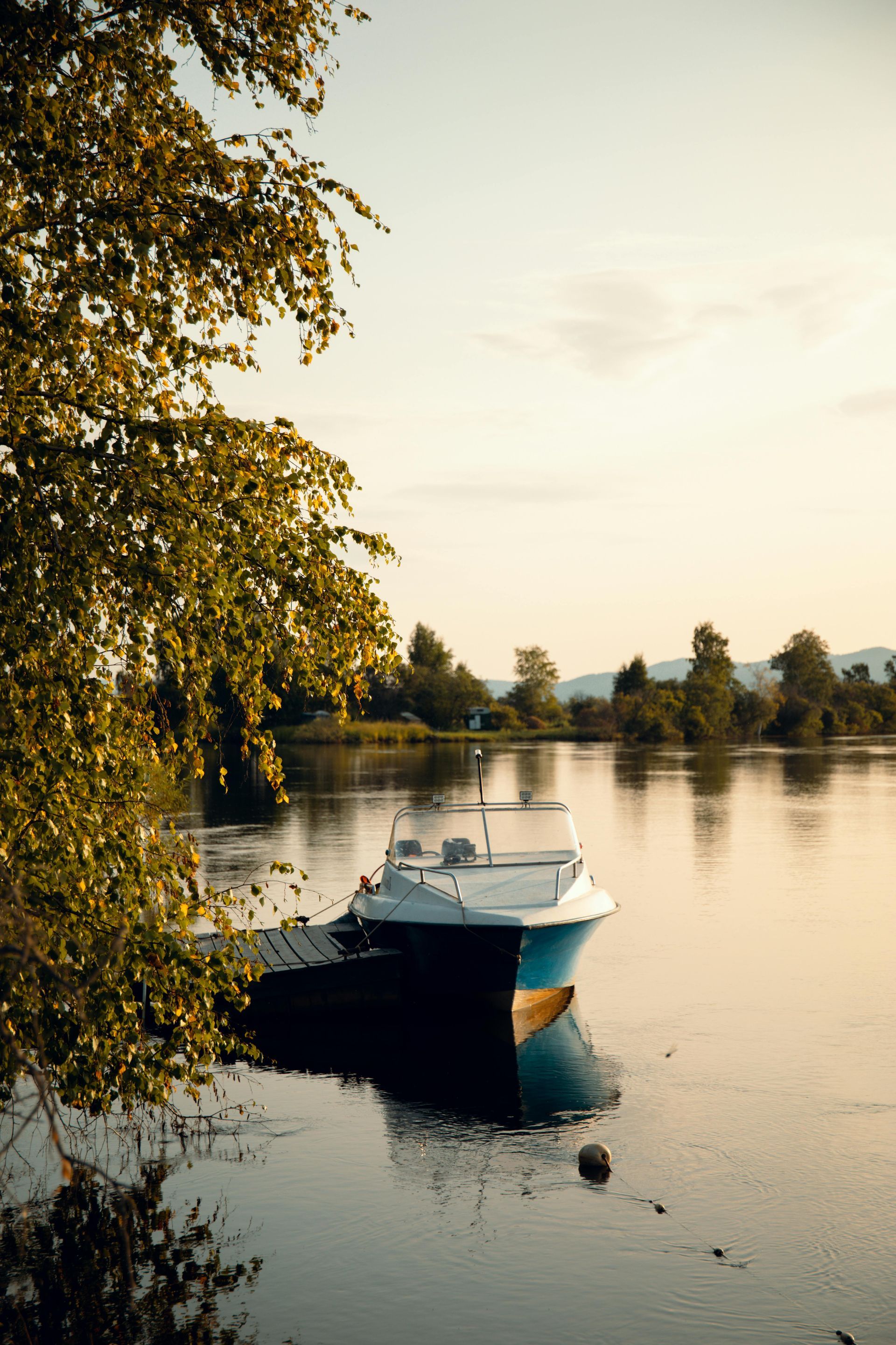 Small white boat moored on a calm lake at sunset, with trees lining the shore and reflecting in the water