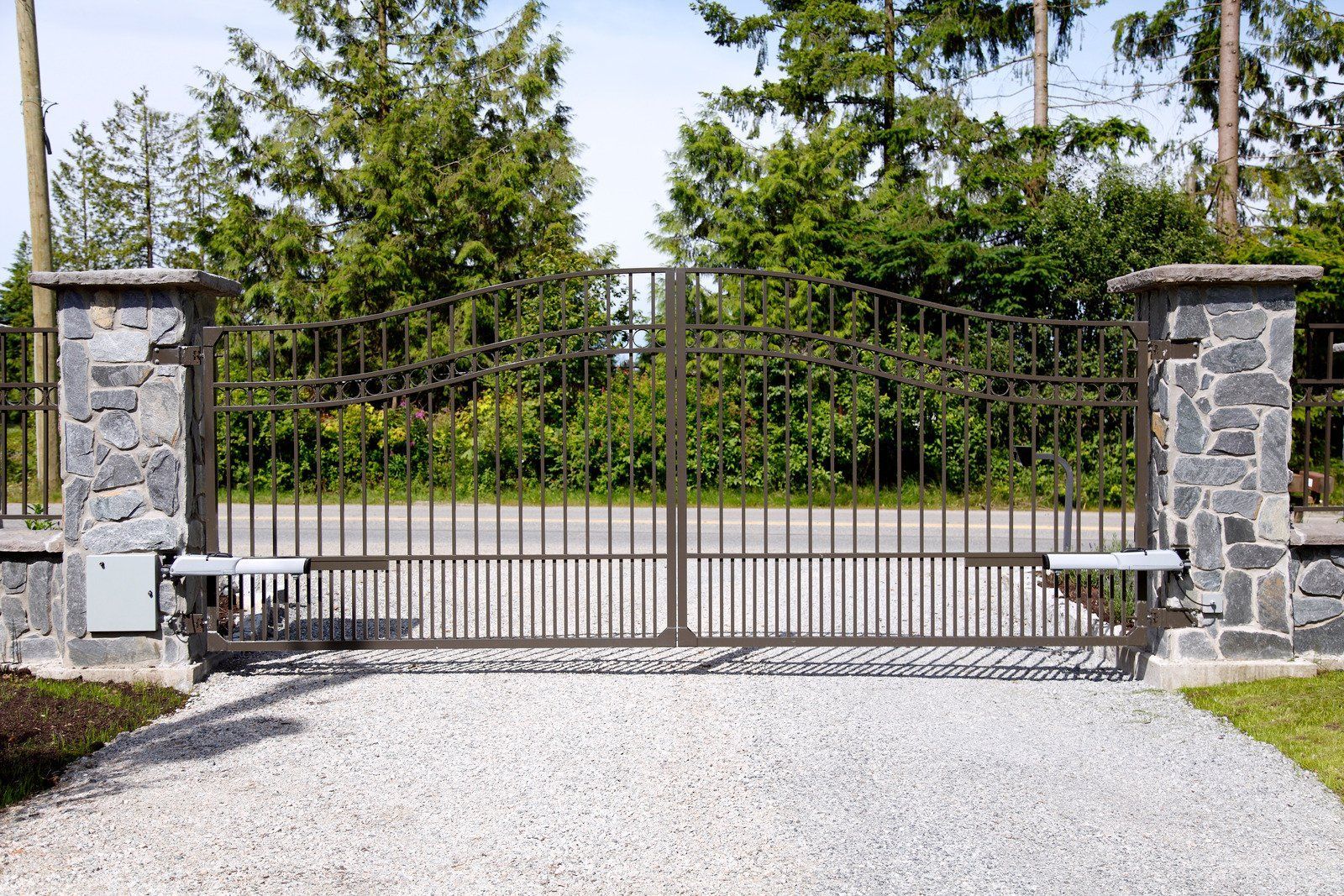Stone gate pillars with a closed metal driveway gate in front of trees and gravel driveway