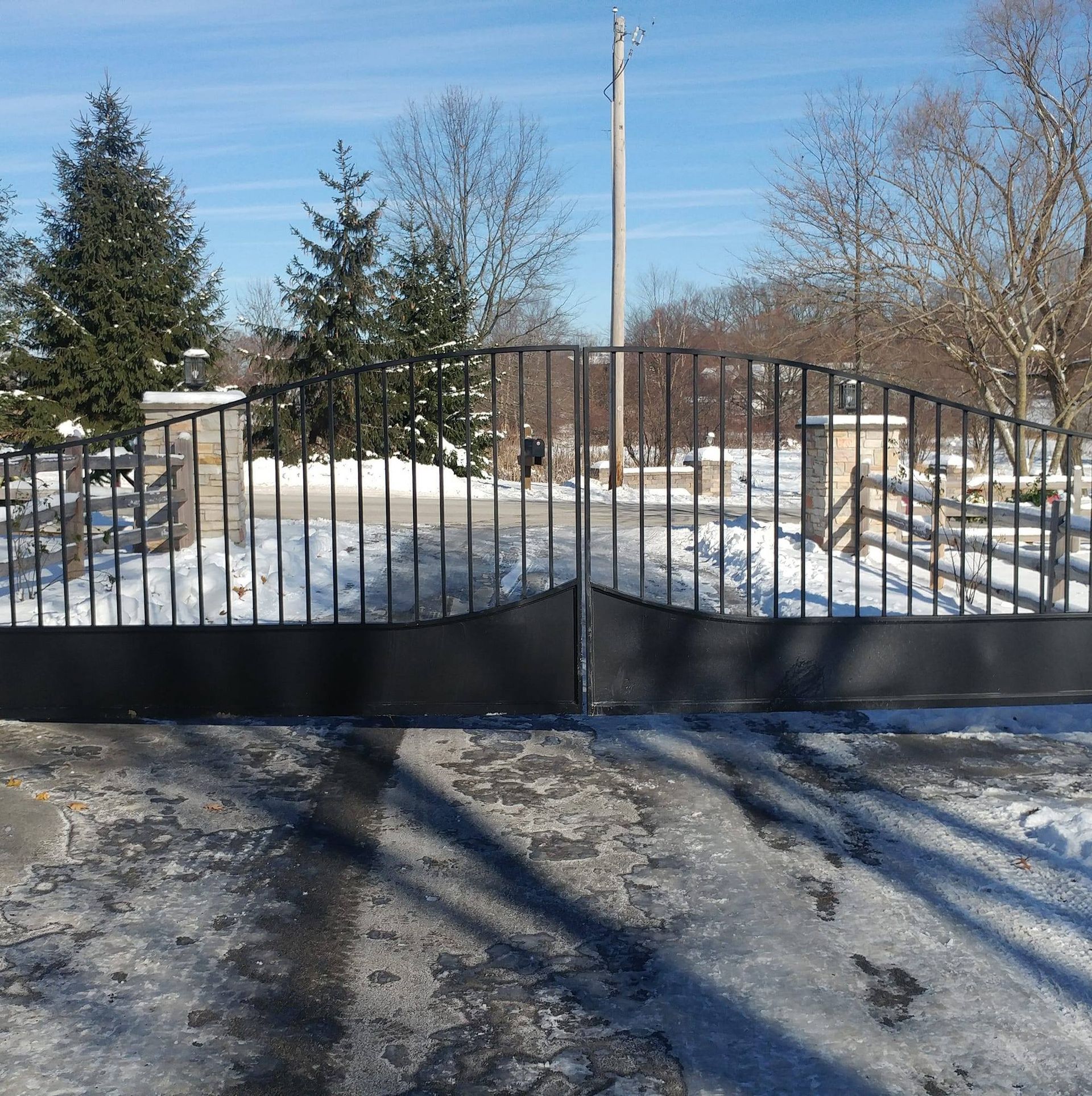 Snowy driveway entrance with black metal gate and trees under a clear blue sky