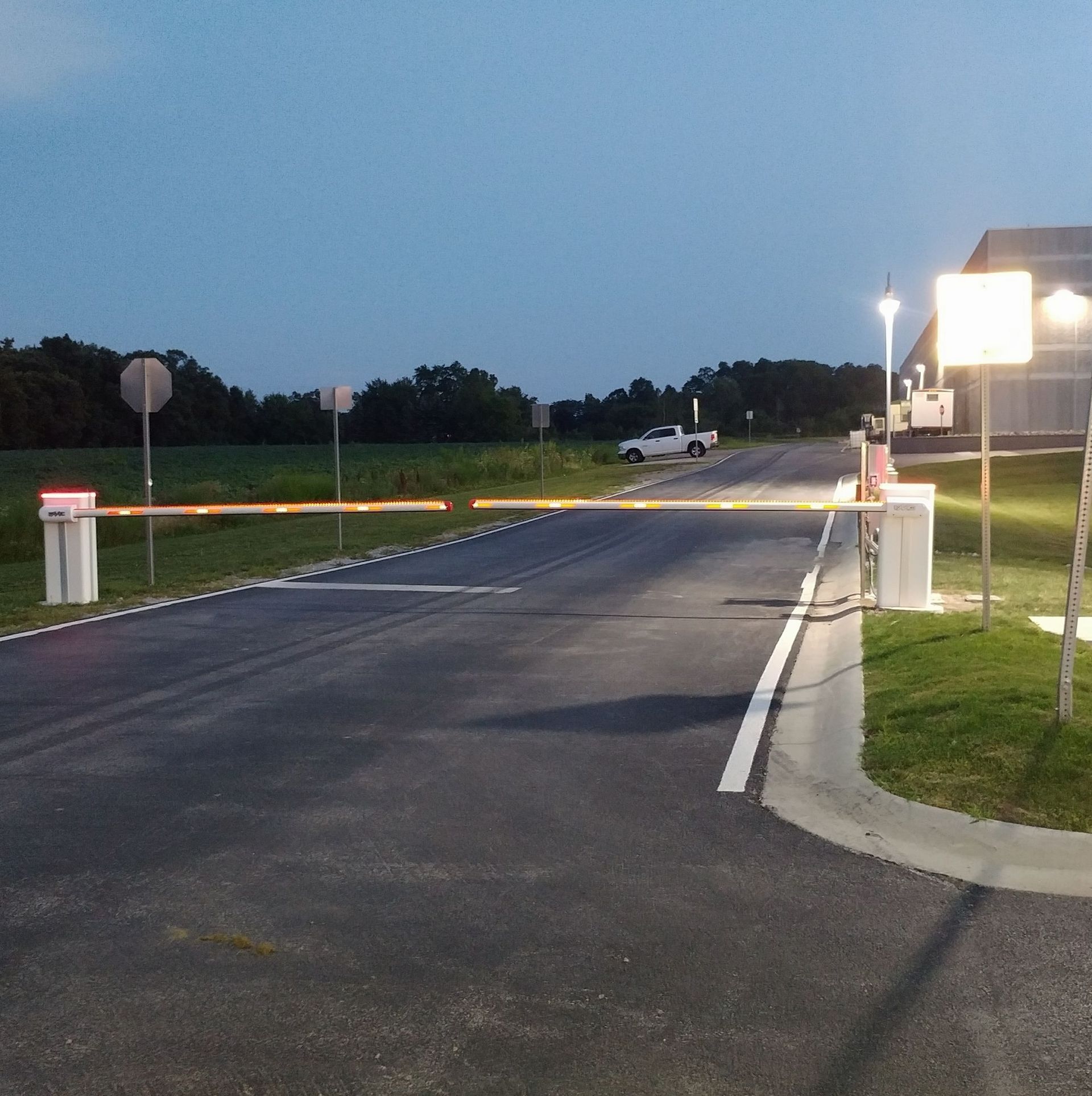 Empty parking lot entrance at dusk with a raised gate, road signs, and a lit building on the right.