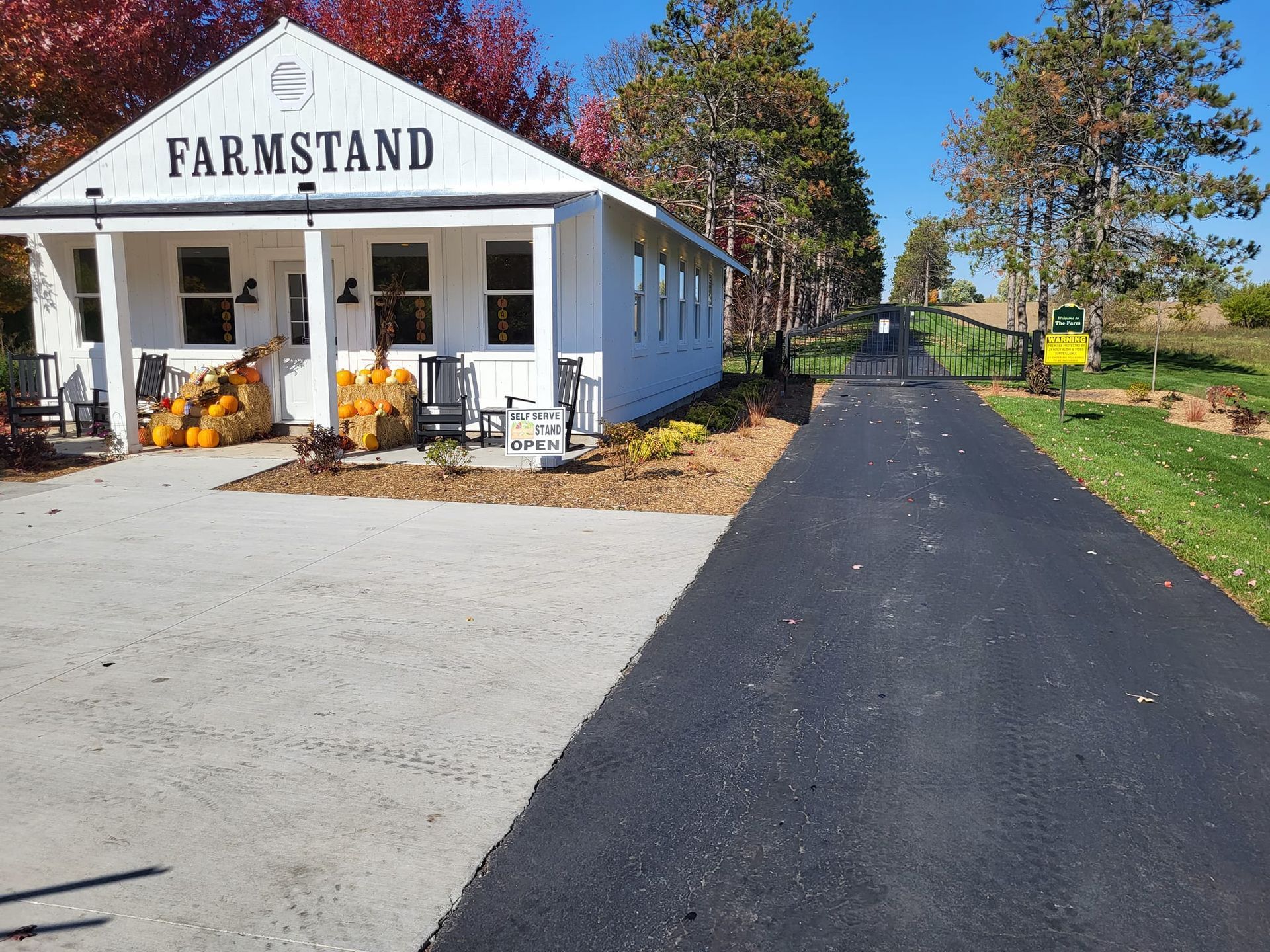 White farmstand with pumpkins by a long paved driveway on a sunny rural property