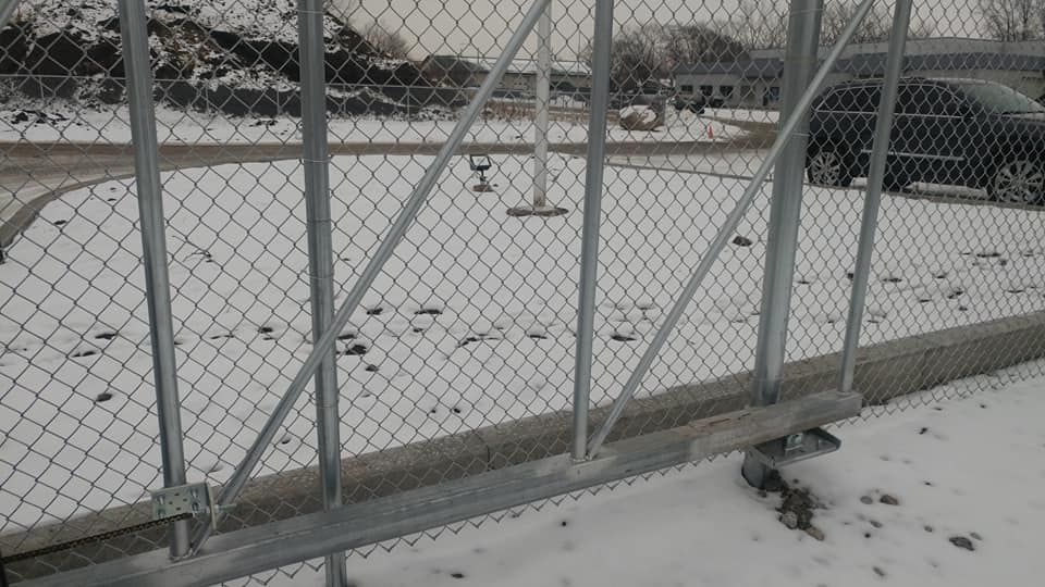 Snow-covered fenced lot with a metal gate and parked vehicles in the background.