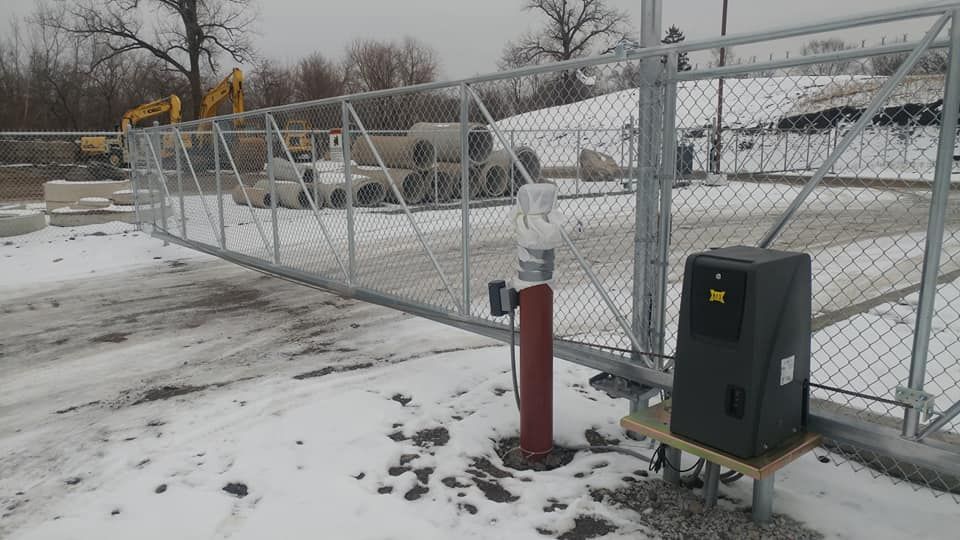 Snow-covered chain-link gate beside red post and gray utility box in an industrial yard
