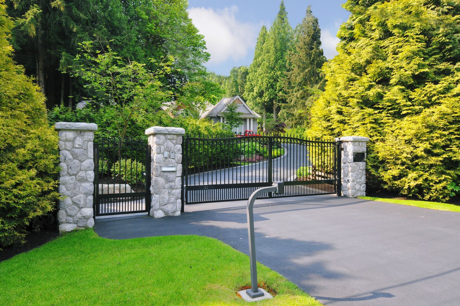Stone gate entrance with black iron gate, flanked by green hedges and trees, beside a paved driveway