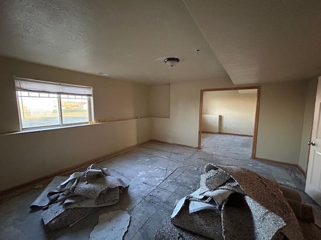 Empty beige room under renovation with exposed subfloor, torn insulation, and a window on the left.