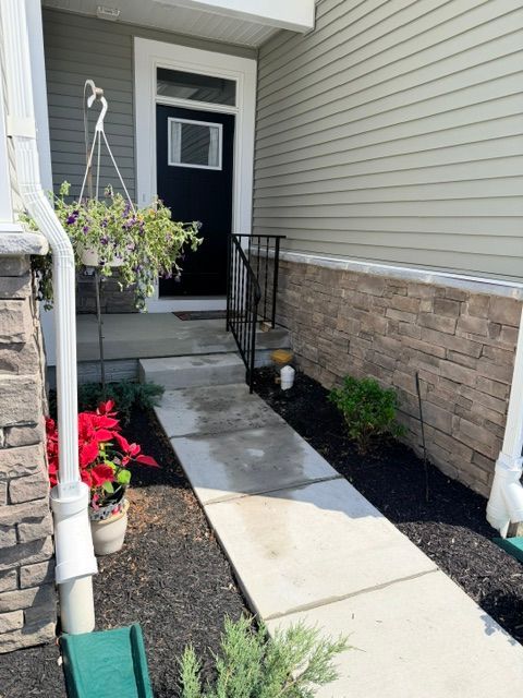 Narrow concrete walkway leading to a front door, with stone siding, hanging flowers, and potted plants.
