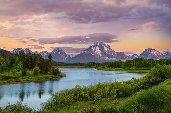 Mountain lake at sunset with snowcapped peaks, green meadow, and calm reflective water