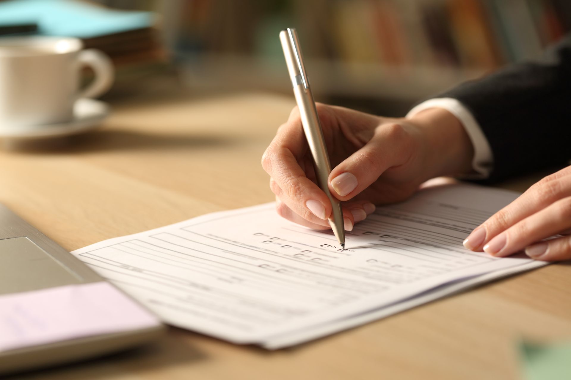 Hand writing on a document at a desk with a pen, beside a cup and laptop.