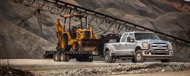 A silver pickup truck towing a trailer with a yellow backhoe in a gravel pit.
