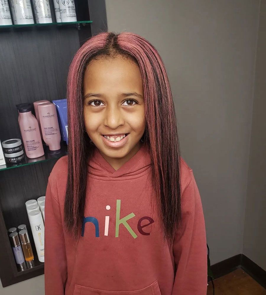 Smiling child in a maroon Nike hoodie with pink braids, standing indoors beside a shelf of hair products