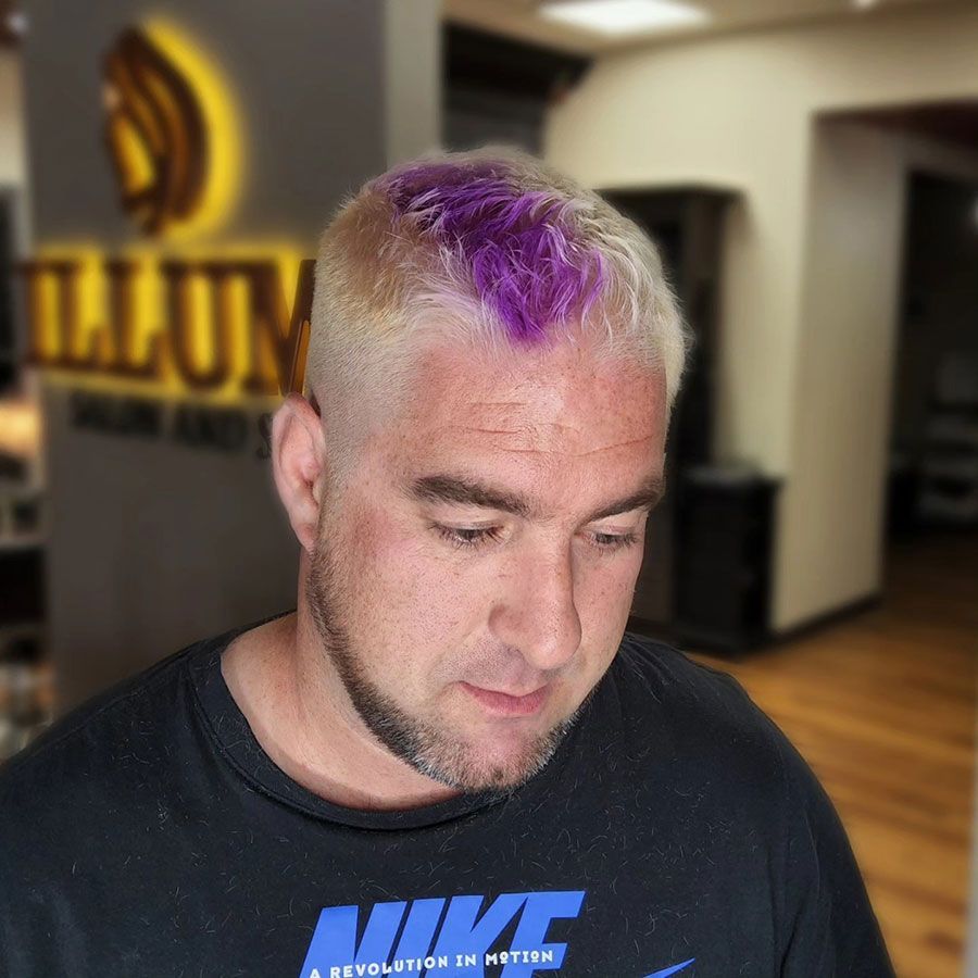 Person with short blond hair and purple-dyed top, looking down in a hallway or lobby.