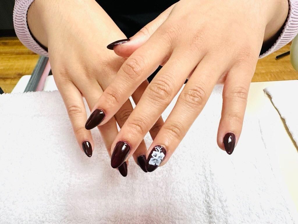 Hands with long dark manicured nails and one white floral accent nail resting on a white table.