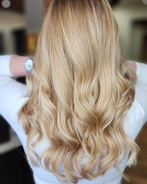 Blonde hair styled in soft loose waves, viewed from behind, with a white top visible at the shoulders