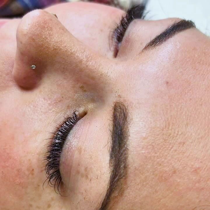 Close-up of a face with eyes closed and a silver nose stud, showing long eyelashes and dark eyebrows.