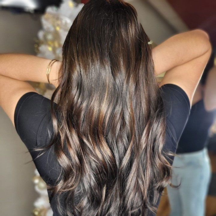 Person with long wavy dark hair seen from behind, arms raised in a blurred indoor setting