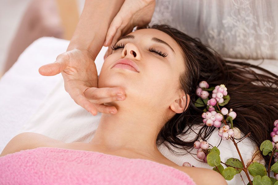 Person receiving a facial massage while lying on a white bed with pink towels and flowers nearby