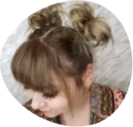 Side view of a woman with a messy blonde bun and bangs, looking downward against a light background.