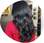 Woman with long wavy black hair wearing a red top, seen from behind in a salon.