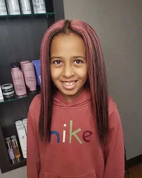 Smiling child in a pink hoodie with long pink braids, standing indoors by a shelf with hair products.