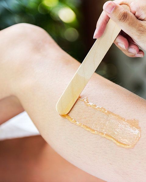 Hand applying wax with a wooden spatula to bare skin on an arm.