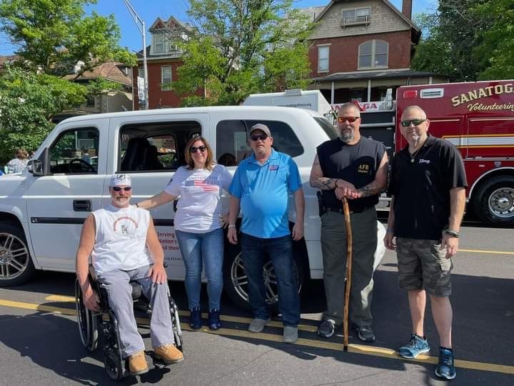 Group of people posing by a white vehicle, with a fire truck in the background, possibly at an event.