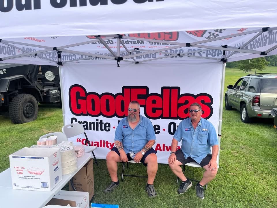 Two men in blue shirts sit in front of a GoodFellas Granite booth. A jeep and SUV are in the background.