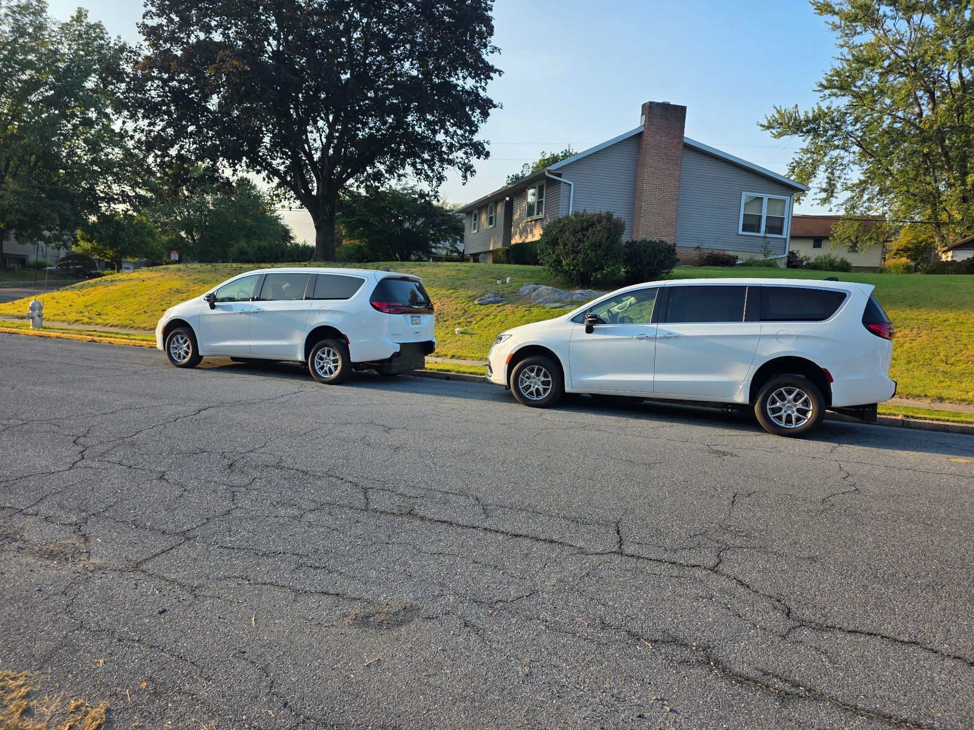 Two white minivans parked on a cracked asphalt street near a house on a sunny day.