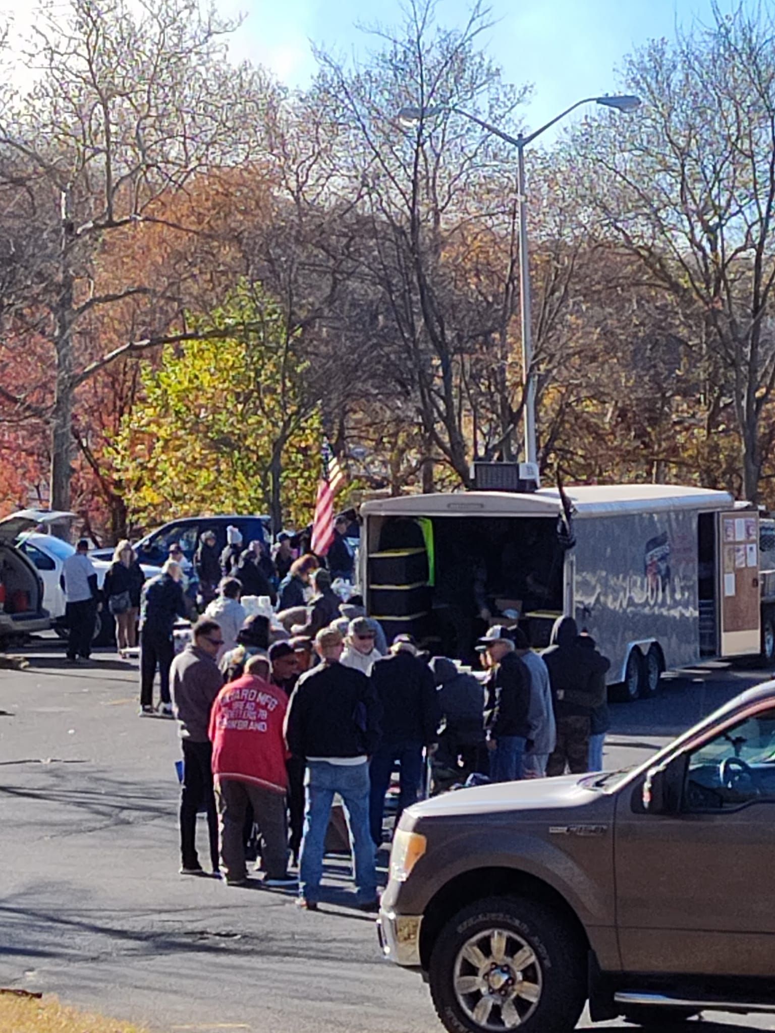 People standing in line near trailers and vehicles; autumn trees in the background.