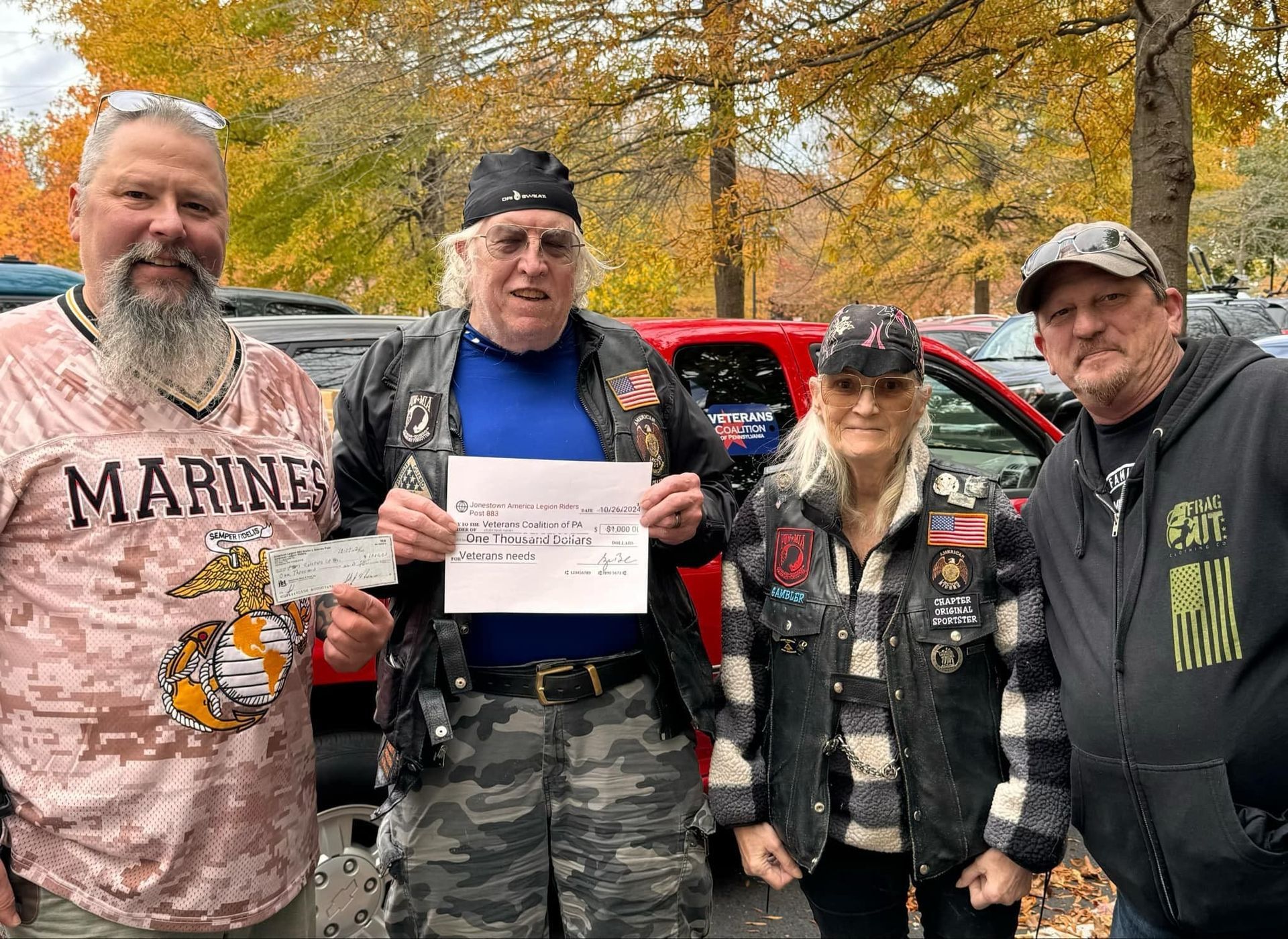 Four people stand outdoors, holding a check. Marines-themed shirt, leather vests, fall foliage.