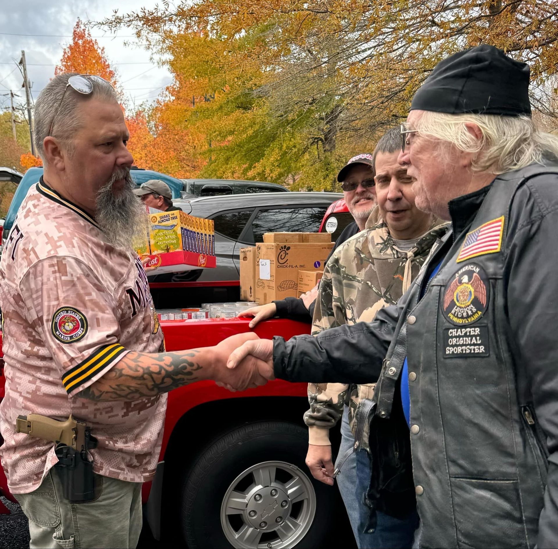 Two men shaking hands near a red truck with boxes, surrounded by others, outdoors.