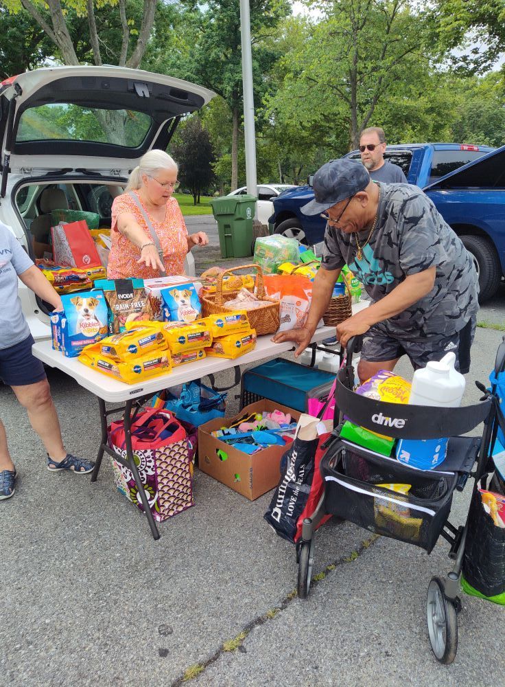 People at a table with goods from a car trunk, outside. A man uses a walker.
