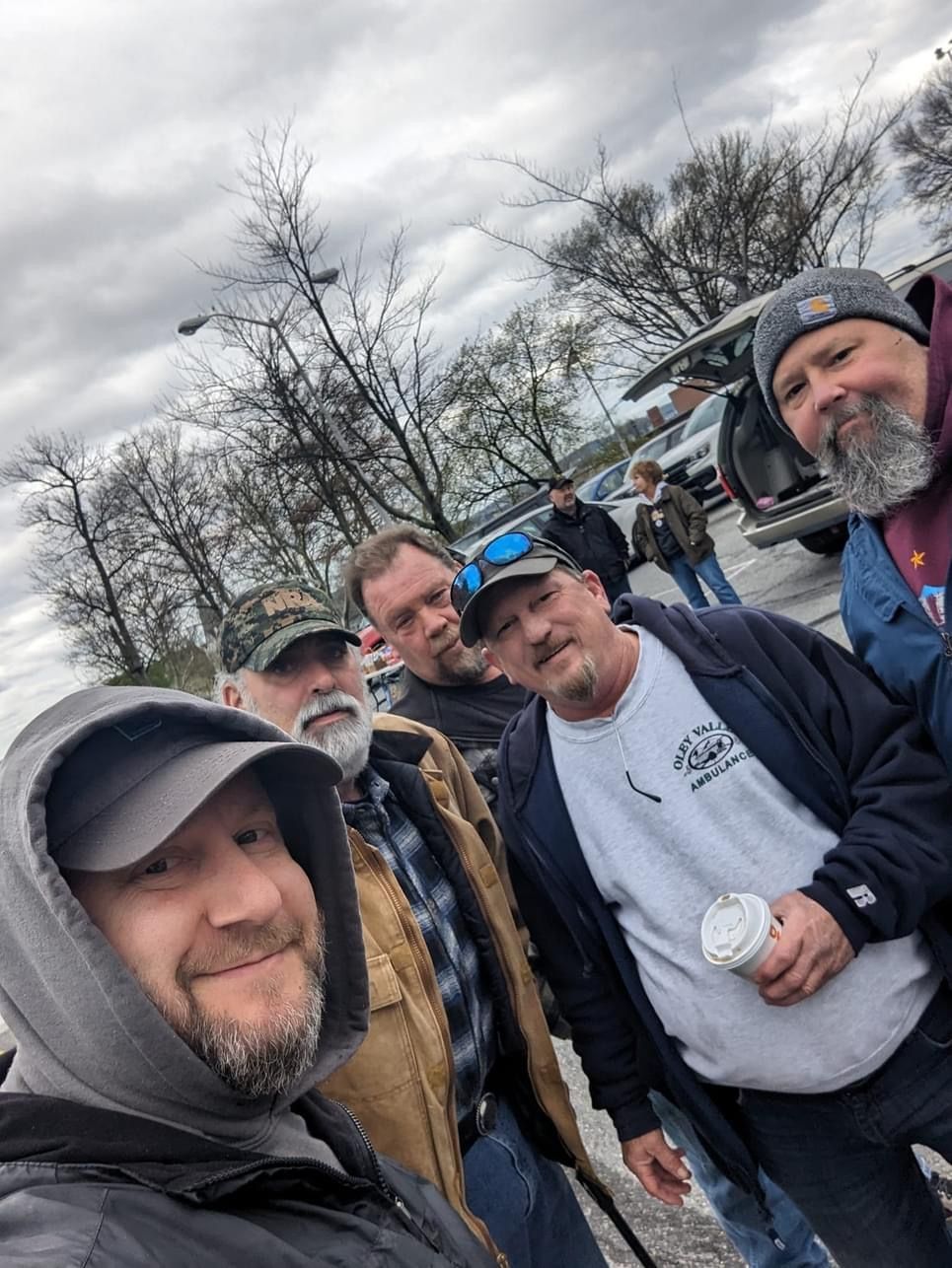 A group of men outdoors, taking a selfie. Overcast sky, some trees, and cars in the background.