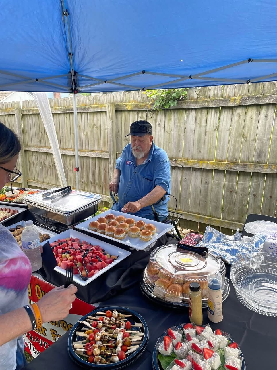 Person behind food table under a blue canopy. Plates of food: skewers, fruit, rolls. A person at left serves.