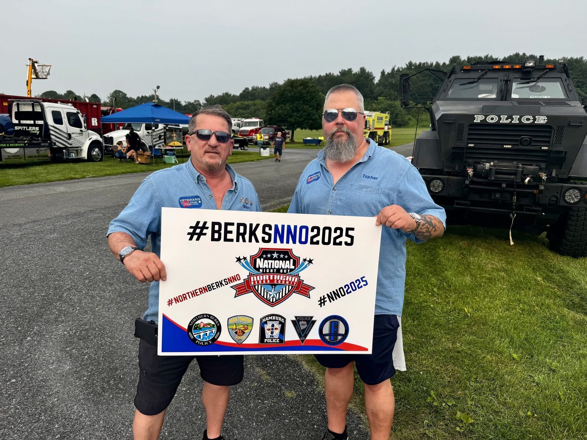Two men holding a sign promoting #BERKSNNO2025. They stand in front of vehicles and a police truck.