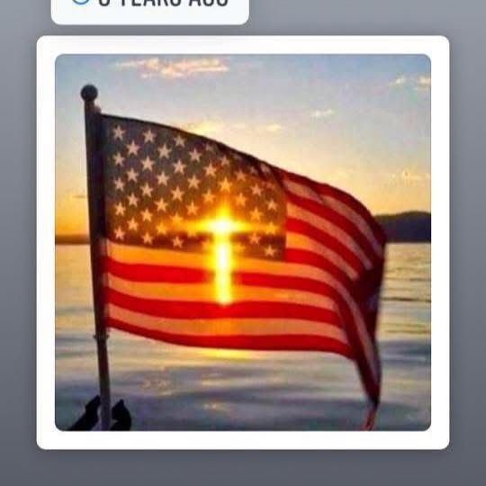 American flag with a cross of light superimposed on it, over water at sunset.