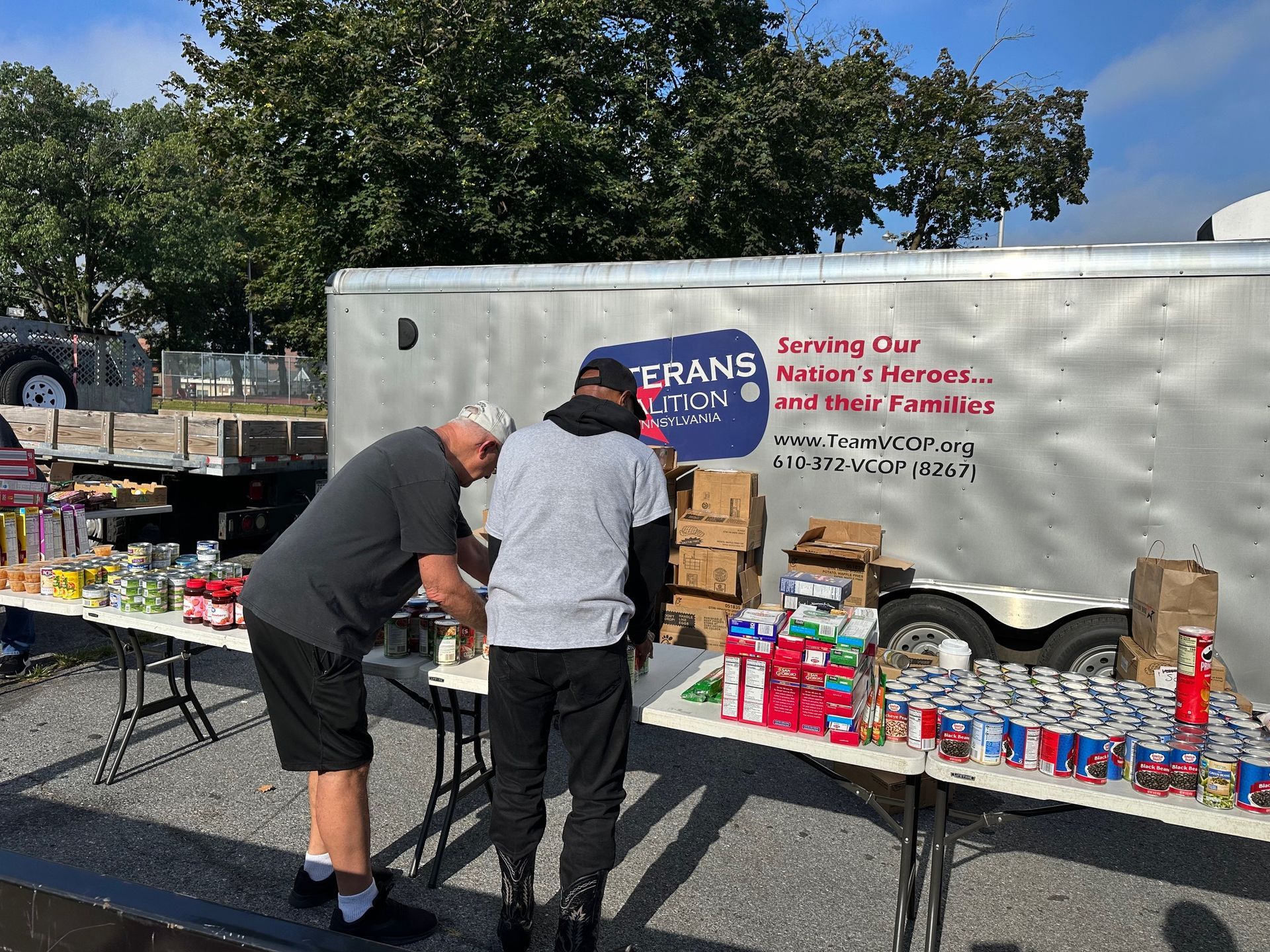 Two people sort food items on tables outside a trailer labeled 