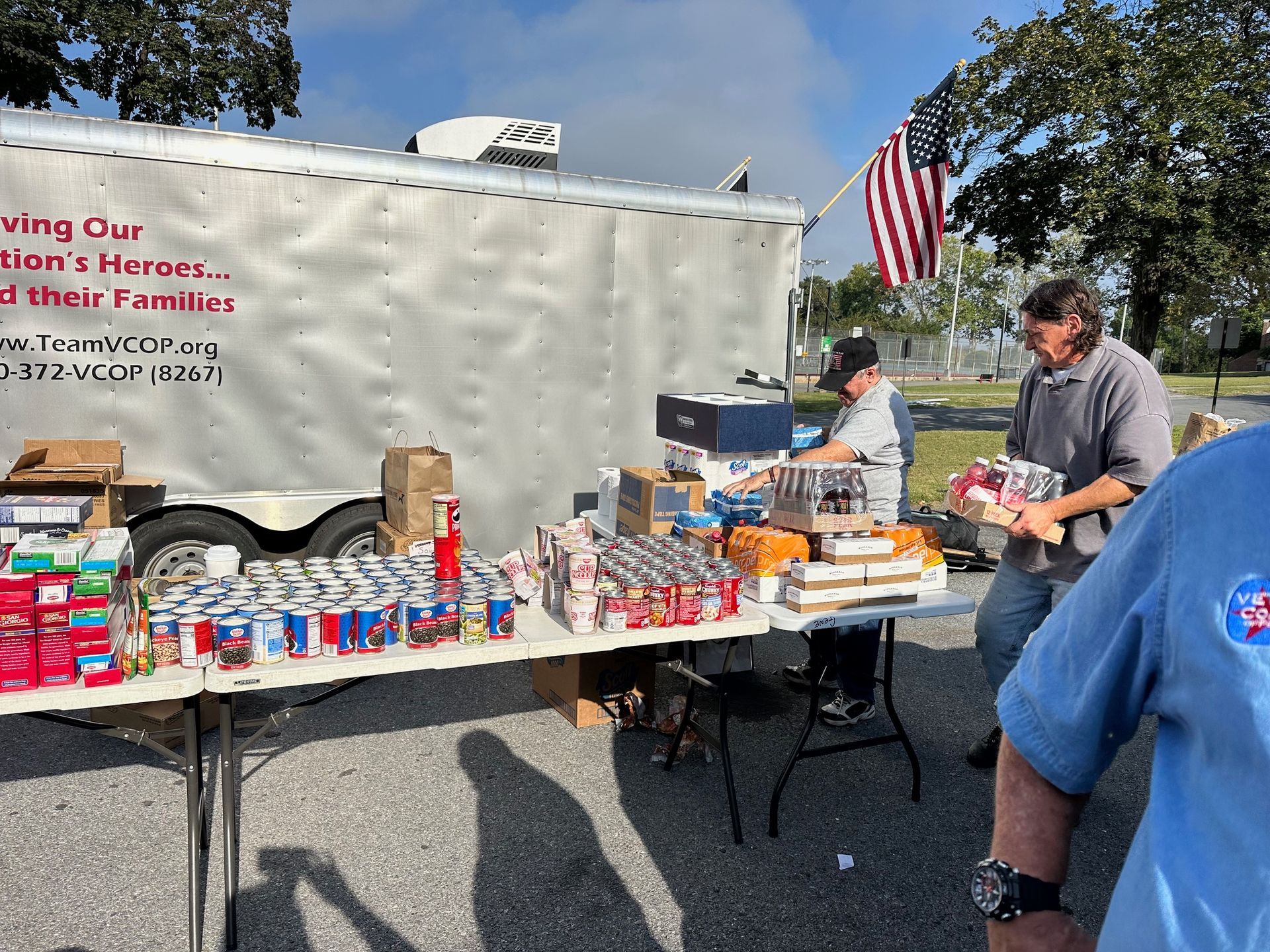 People distributing food from a trailer with an American flag. Canned goods and packaged food are on tables outside.