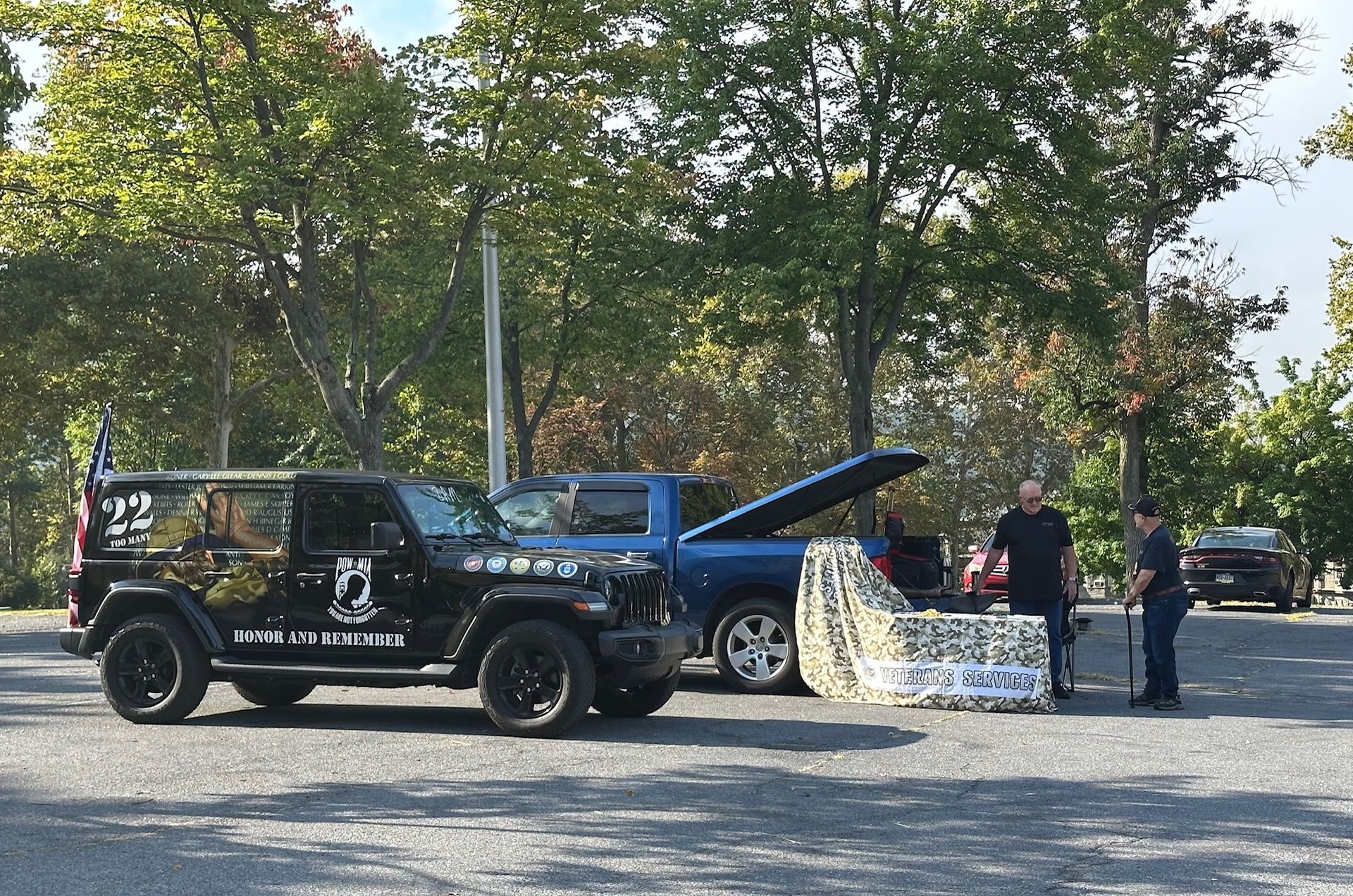 Black Jeep and blue truck in a parking lot; people stand near. Trees in background, sunny day.