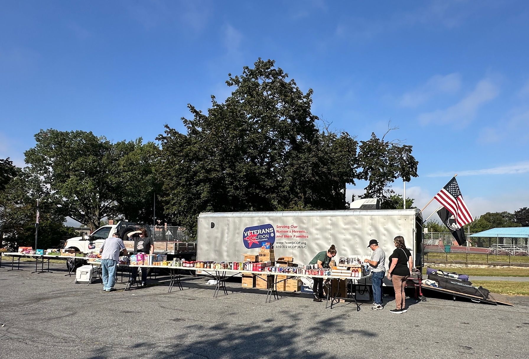 Outdoor vendor setup with tables displaying merchandise. A trailer sits behind tables, an American flag waving nearby.