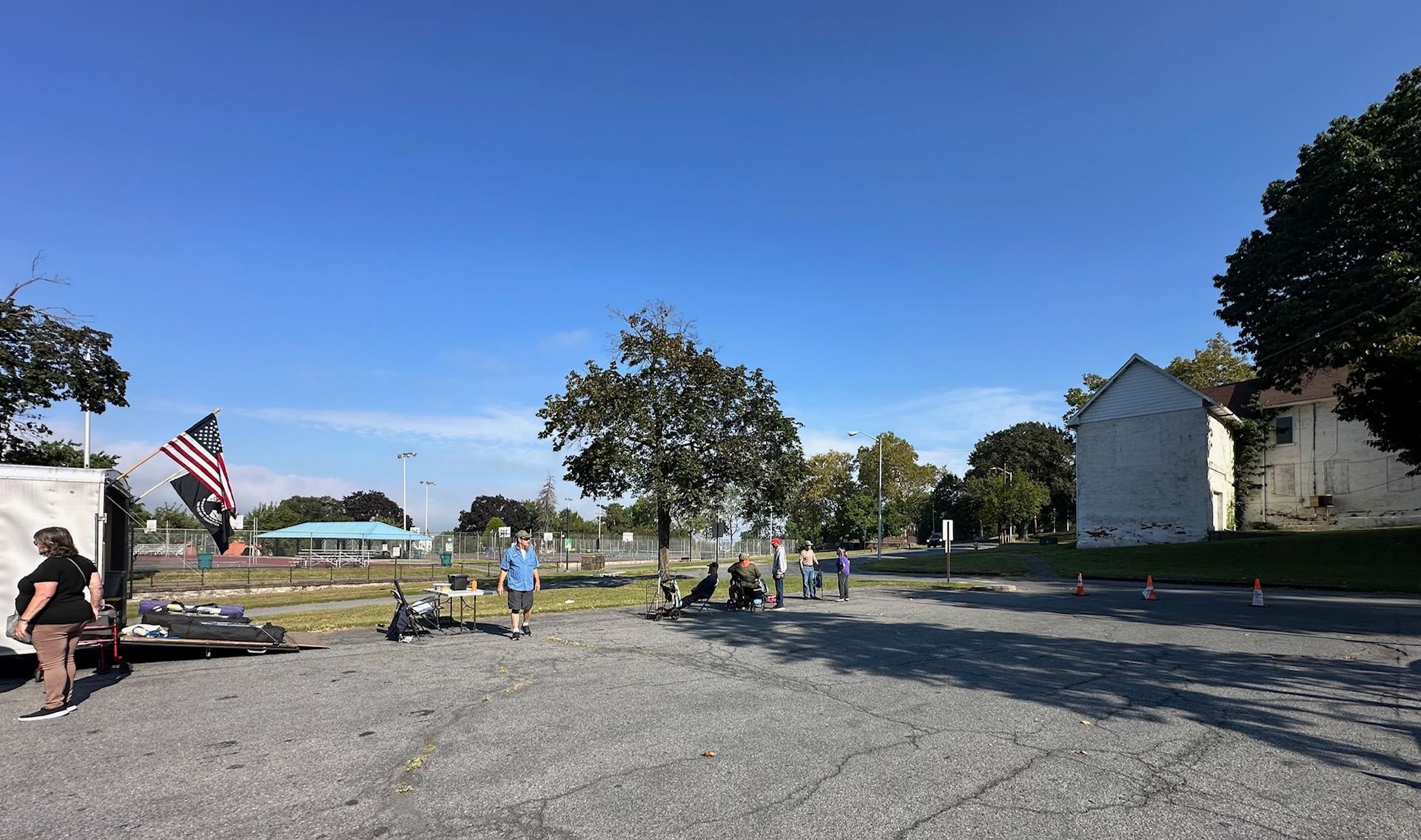 Gravel parking lot on a sunny day. People near a tree and building. American flag visible.